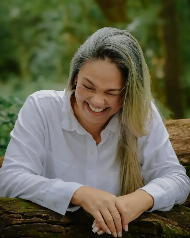 A Dinda, responsável pelo ateliê, rindo apoiada em um tronco ao ar livre