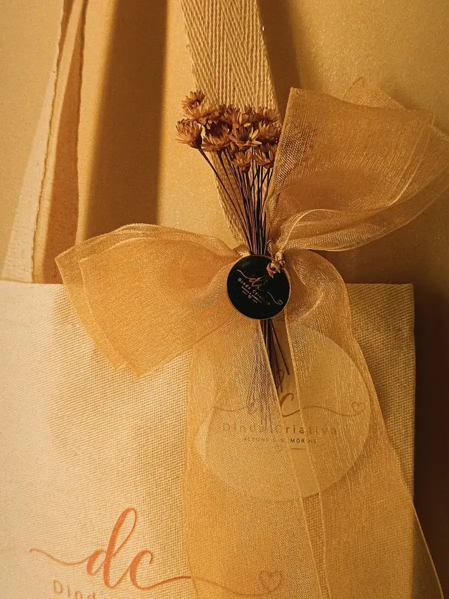 Álbum embalado para presente em sacola de tecido, com laço de organza dourado, flores secas e tag da marca Dinda Criativa